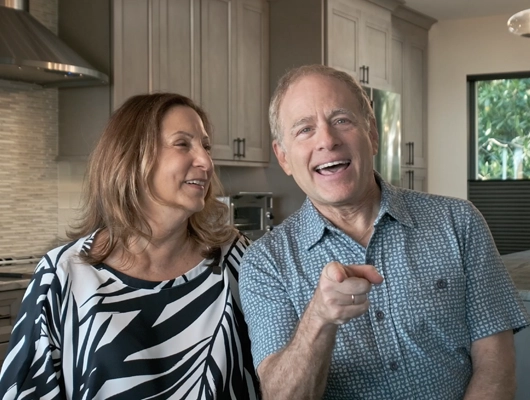 man pointing at camera sitting next to woman in kitchen